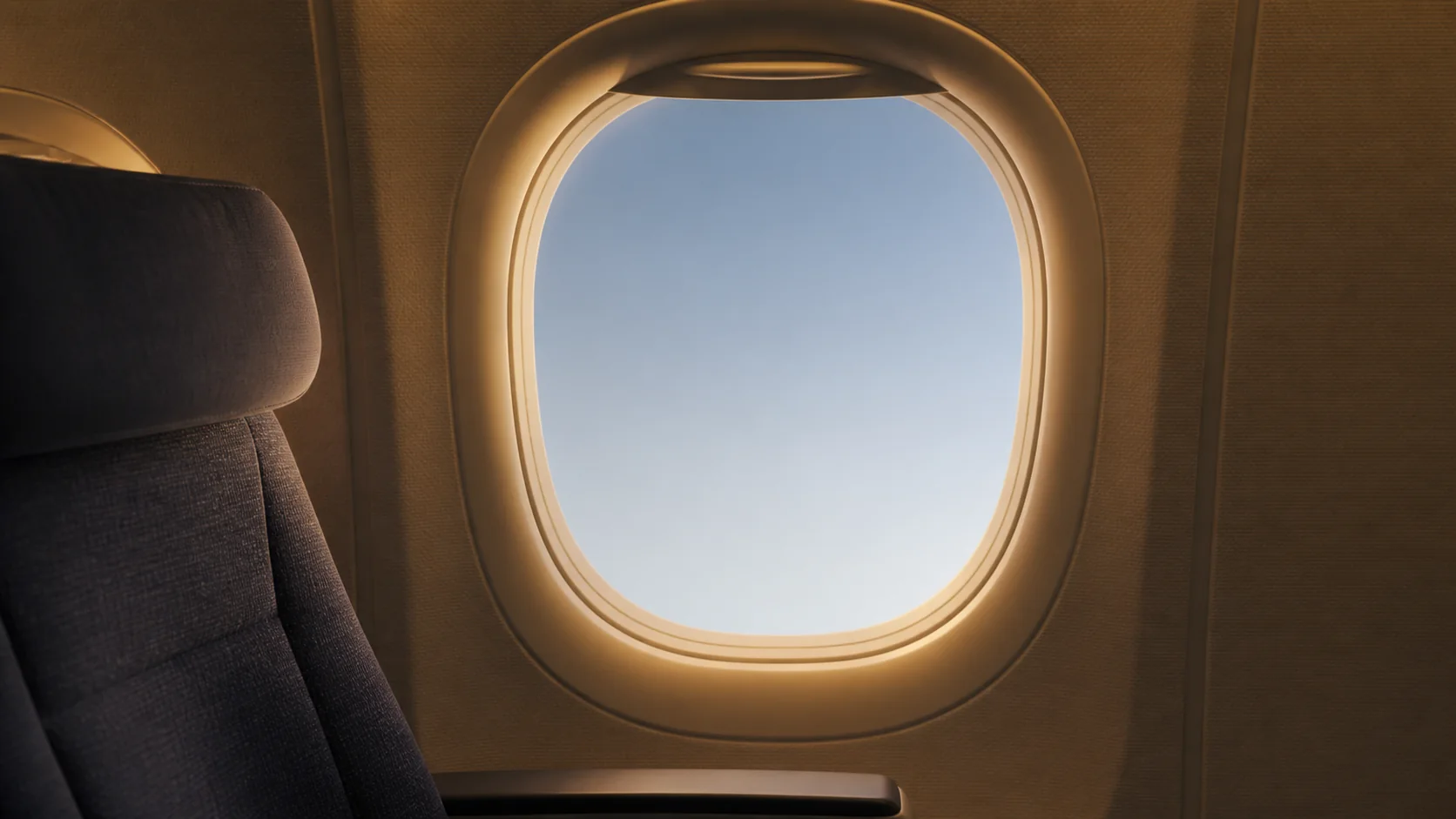 An empty airplane window seat overlooking softly lit clouds during a calm golden-blue hour flight